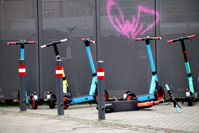 Blue and black kick scooters lined up at a bike rack in front of a perforated metal wall with pink graffiti.